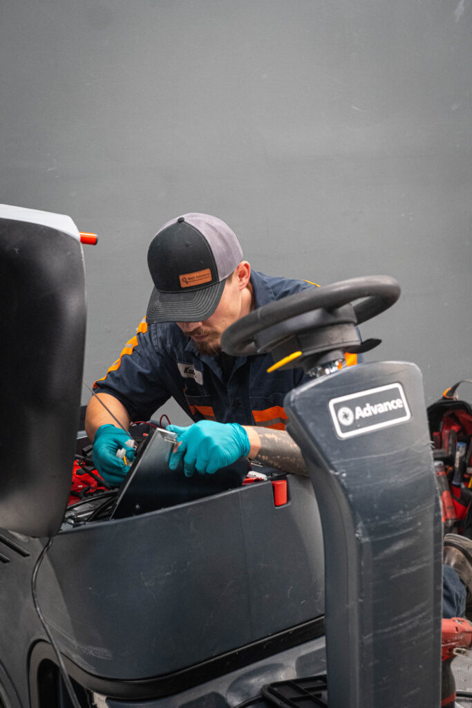 WaySolutions technician Kaden inspecting the internal electrical wiring and battery compartment of an Advance ride-on floor scrubber.
