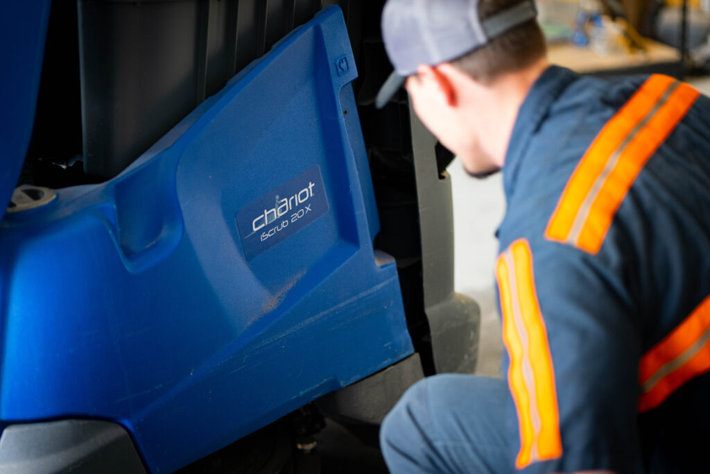 A WaySolutions technician kneels to inspect the lower housing of a blue Kärcher Chariot iScrub 20X floor scrubber in Layton, Utah.
